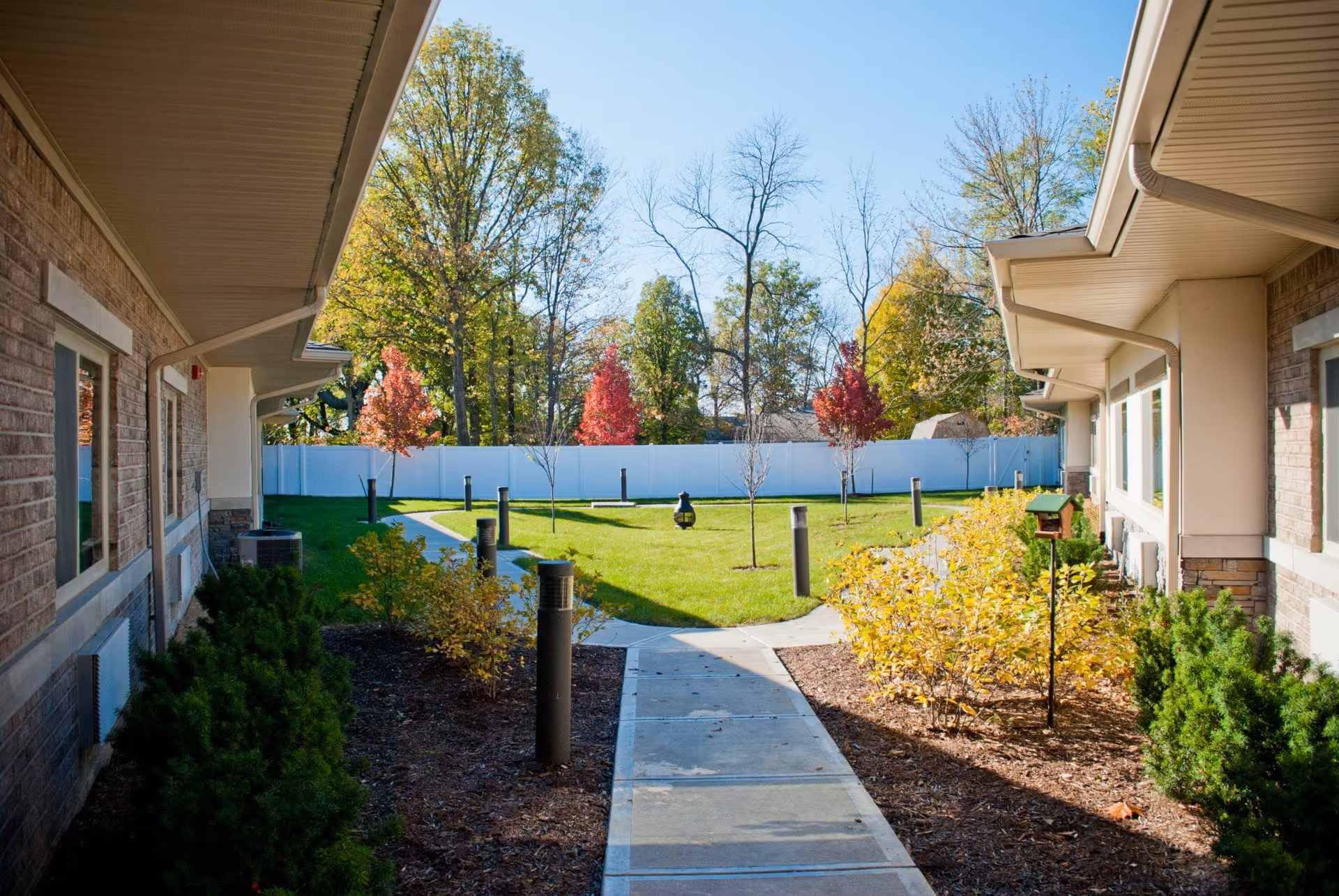 A sunny outdoor courtyard area between two brick buildings with a concrete walkway leading to a grassy area with small trees and shrubs. The courtyard is surrounded by a white fence and tall trees with autumn foliage in the background.