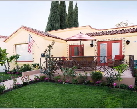 Exterior view of a single-story building with a tiled roof and beige walls, featuring a small patio area with a red and white striped umbrella, outdoor seating, and decorative plants in front. An American flag is mounted near a window, and tall evergreen trees are visible in the background.