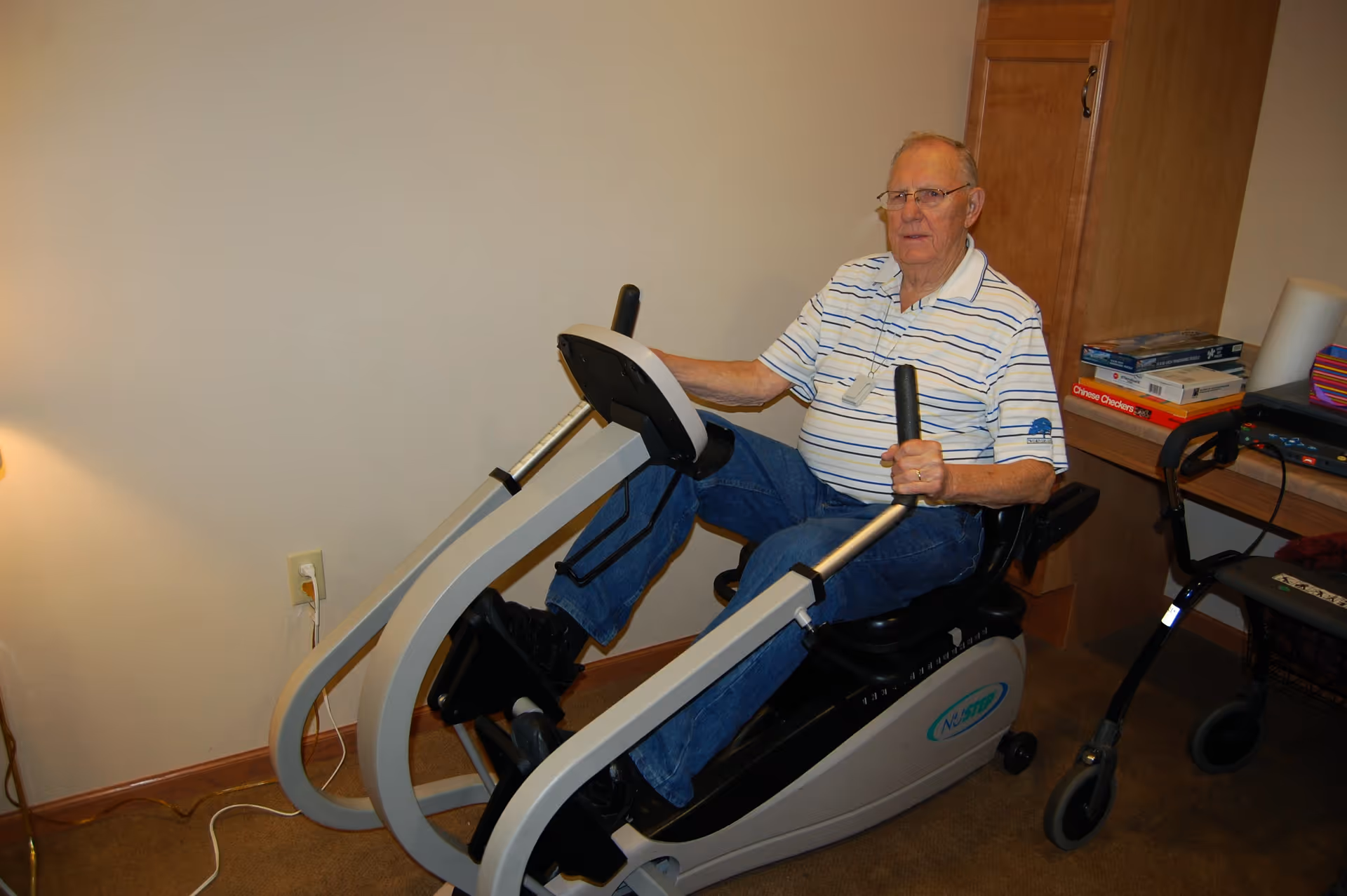 An elderly man wearing glasses, a striped polo shirt, and jeans is seated on a recumbent exercise bike in a room with beige walls and carpet. Behind him is a wooden cabinet with books and other items on top, and a walker is positioned nearby.