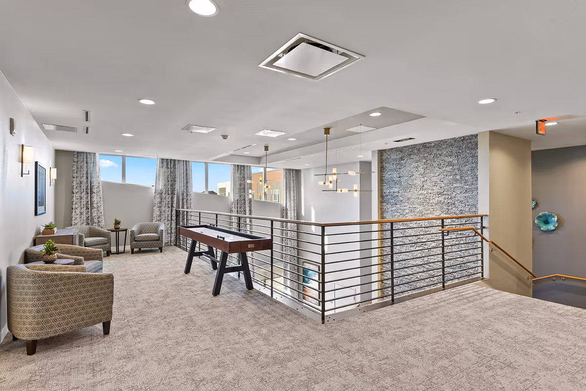 A spacious, carpeted lounge area with modern armchairs and small tables along the left wall. A shuffleboard table is positioned near the railing overlooking a lower level. Large windows with patterned curtains allow natural light to fill the space. Contemporary pendant lights hang from the ceiling, and a textured stone accent wall is visible near a staircase with wooden handrails.