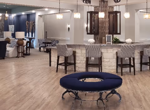 Spacious interior of a senior living facility featuring a circular blue cushioned bench in the foreground, a stone-faced bar counter with four high chairs, pendant lights hanging above, and a seating area with sofas and tables in the background.
