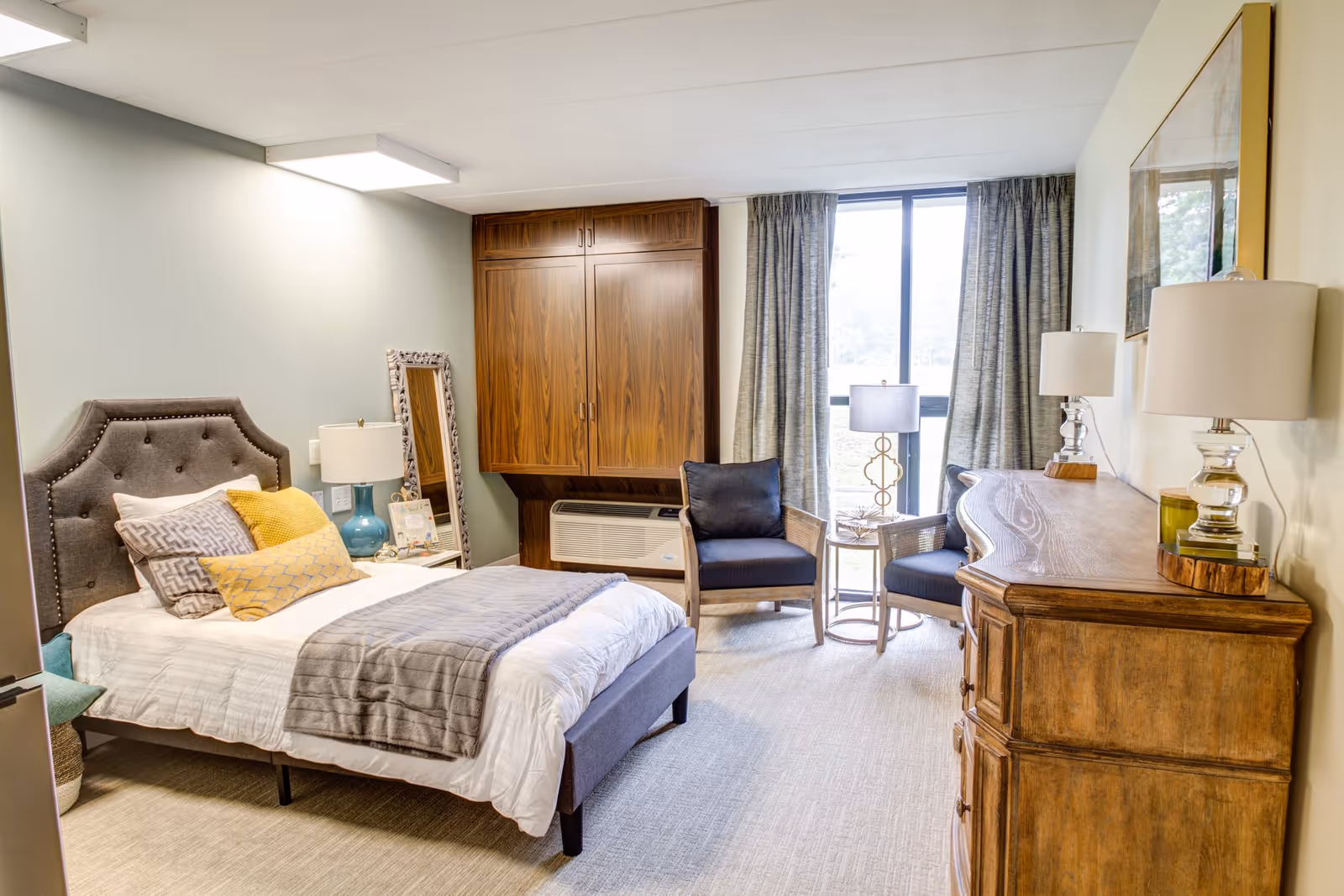 A cozy bedroom with a gray upholstered bed featuring multiple pillows and a gray blanket. There is a wooden dresser with two lamps on top, two cushioned chairs with a small round table between them near a large window with curtains. A wooden cabinet is mounted on the wall above an air conditioning unit, and a decorative mirror leans against the wall next to the bed.