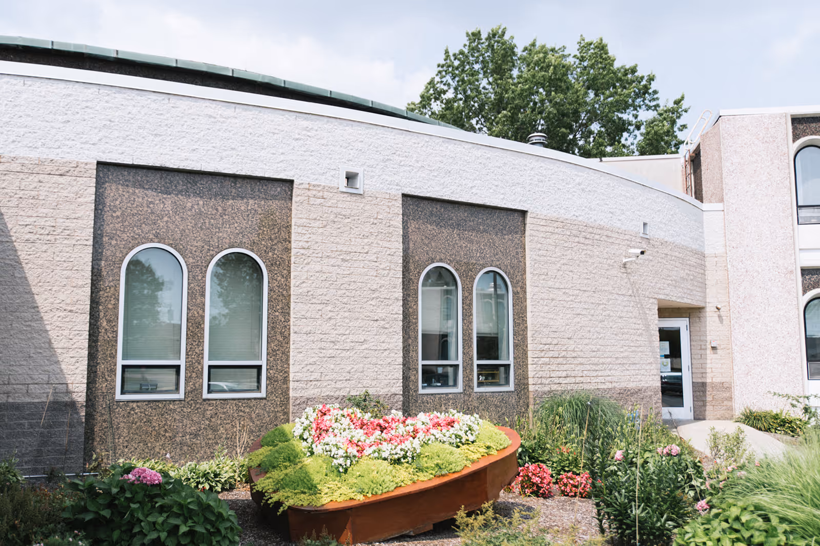 Exterior view of a building with beige and brown textured walls, featuring four arched windows. In front of the building, there is a garden with various green plants and colorful flowers arranged in a raised, boat-shaped planter. Trees and a partly cloudy sky are visible in the background.