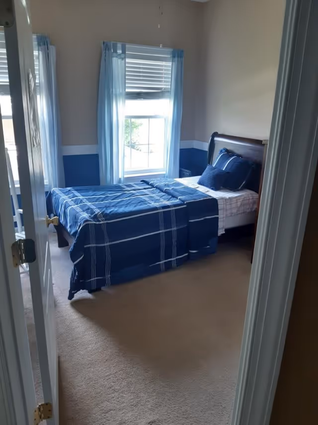View through an open door into a bedroom with a single bed made with blue and white plaid bedding. The room has beige walls with a blue and white chair rail, two windows with light blue curtains, and beige carpeted floor.