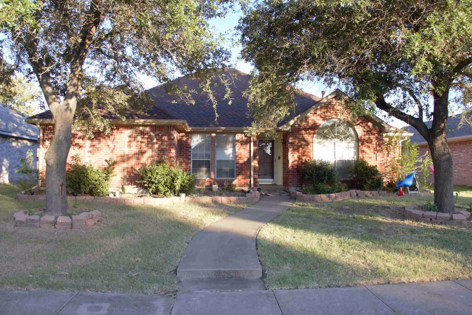 Front exterior view of a single-story brick house with a curved concrete walkway leading to the front door. The house has two large windows, one with an arched top, and is surrounded by green grass, bushes, and two large trees in the front yard.