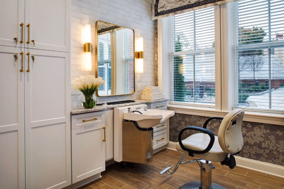 A bright and clean salon area with a white styling chair in front of a sink and a large mirror mounted on a white brick wall. The countertop has a vase of white flowers and a stack of towels. Large windows with blinds allow natural light to fill the room, showing an outdoor view.