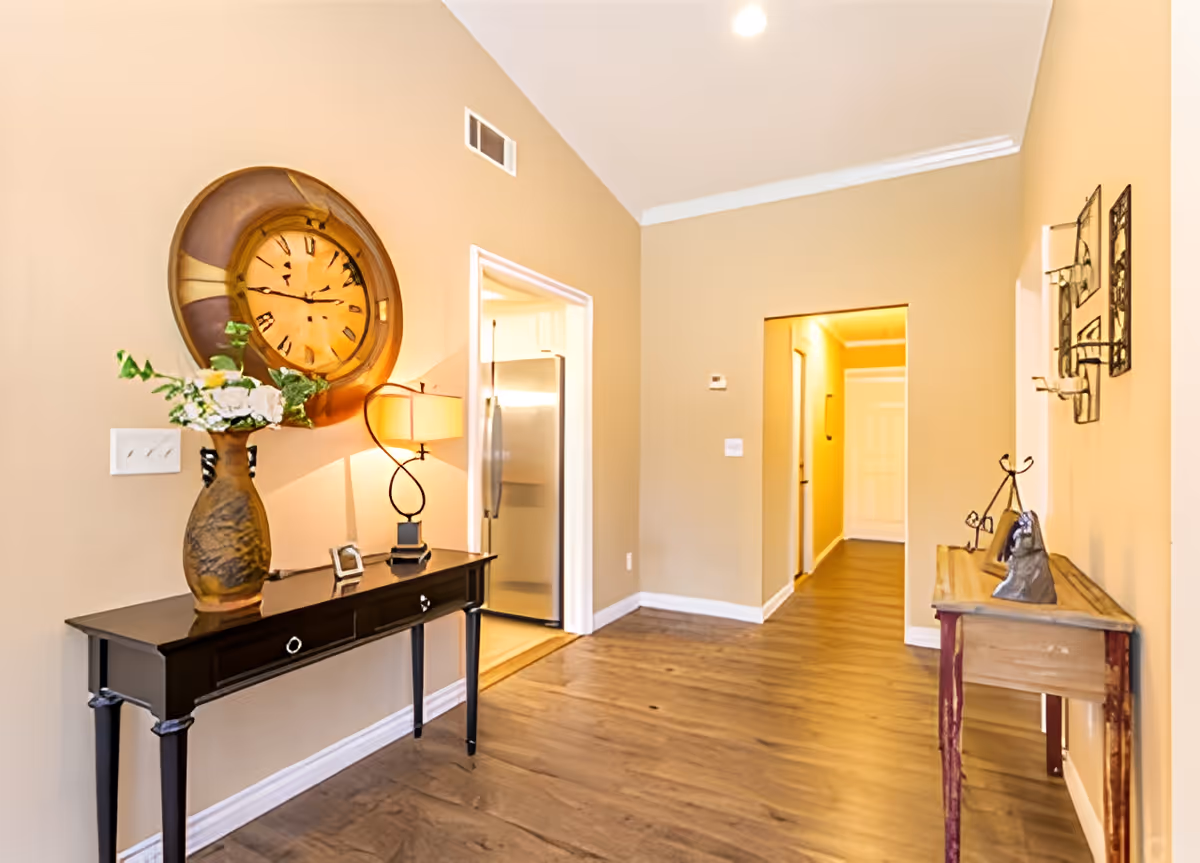 Interior hallway of a senior living facility with wooden flooring and beige walls. On the left, there is a black console table with a decorative vase holding flowers and a lamp. Above the table, a large round wall clock is mounted. On the right side, there is a wooden side table with decorative items and wall-mounted candle holders. A doorway leads to a kitchen area with a stainless steel refrigerator, and another hallway extends further into the building.