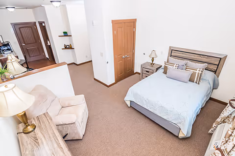A cozy bedroom in a senior living facility featuring a bed with pillows and a light blue bedspread, a wooden nightstand with a lamp, a comfortable beige armchair, a wooden dresser with a lamp, and a closet with double doors. The room has beige carpet and white walls with wooden trim.
