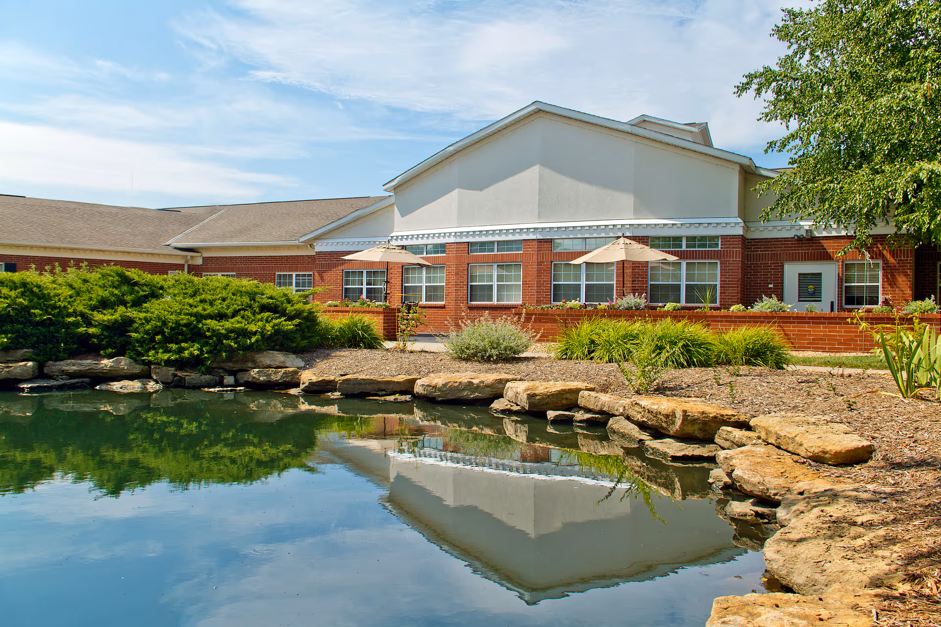 Brick and stucco senior living building with patio umbrellas, landscaping, and a reflecting pond in front.
