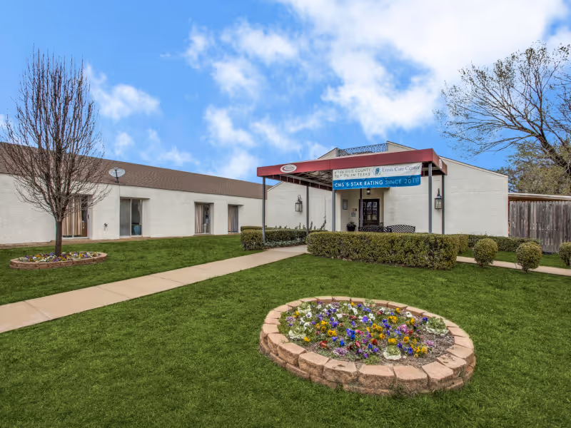 Exterior view of Ennis Care Center showing a single-story building with a covered entrance. There is a well-maintained lawn with flower beds and small trees, and a clear blue sky with some clouds above.