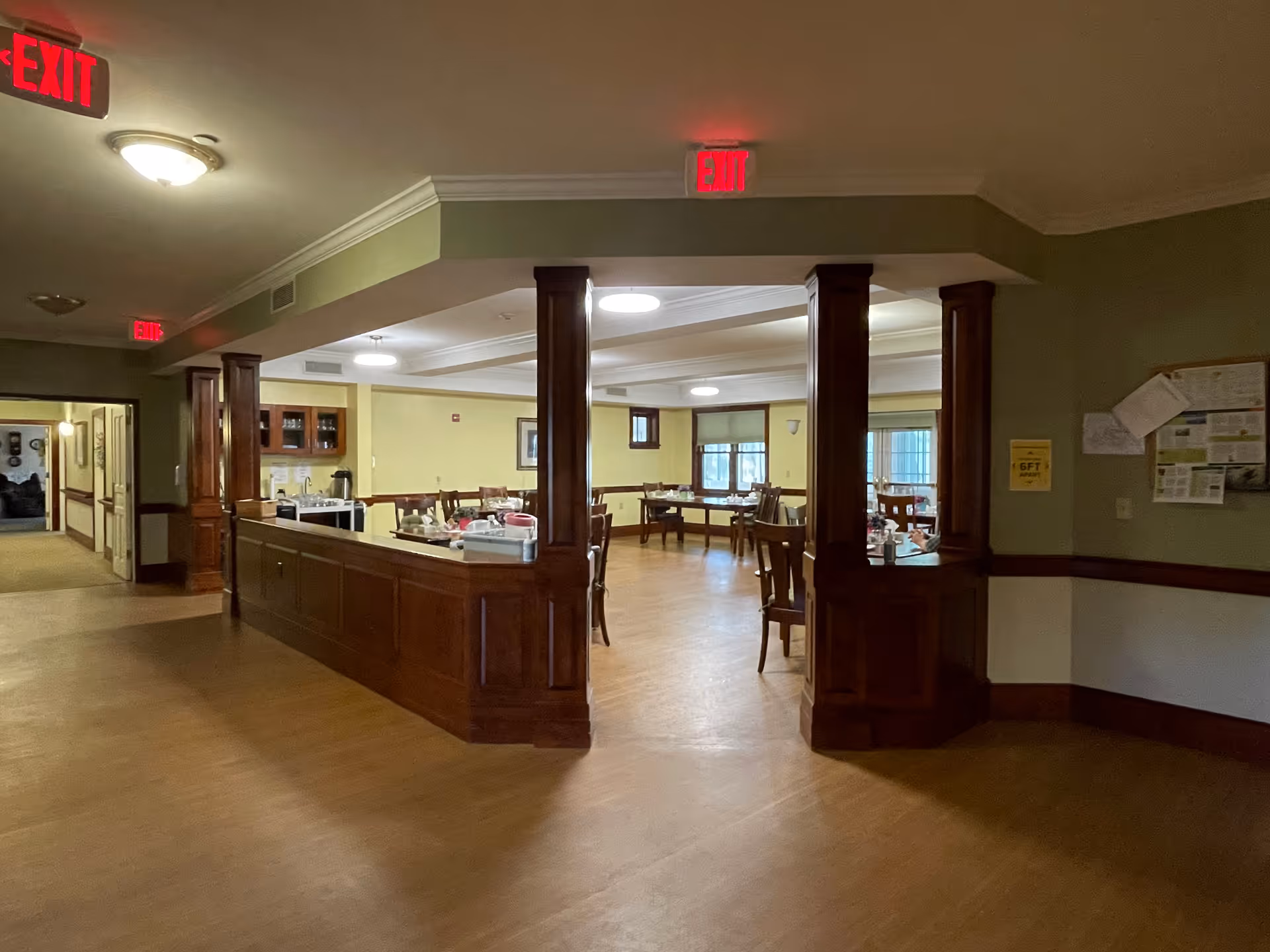 Interior view of a senior living facility dining area with wooden chairs and tables set for meals. The room has soft yellow walls, wooden trim, and multiple ceiling lights. There are exit signs illuminated in red above doorways and a hallway leading to other rooms.