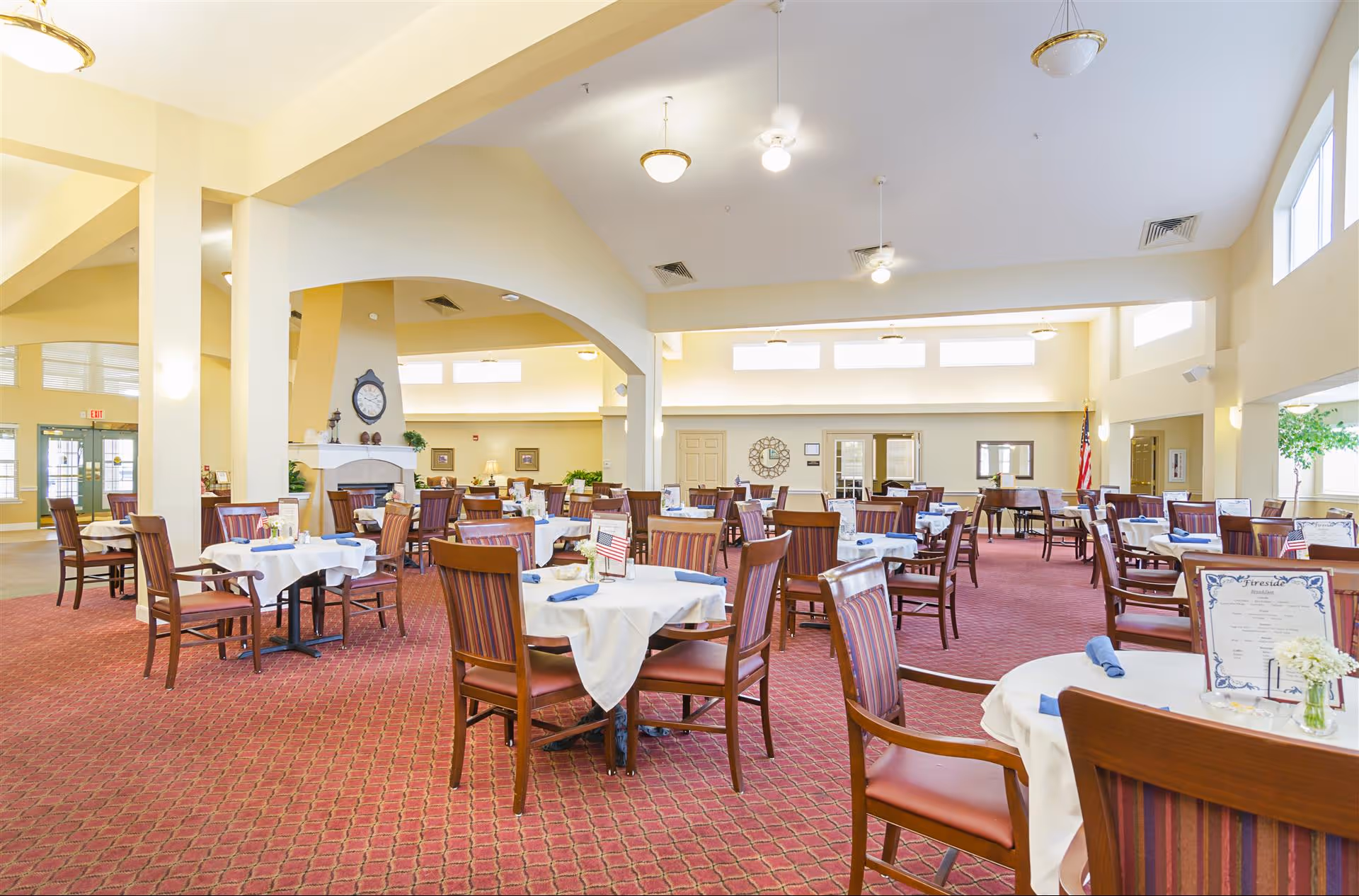 A spacious dining room with multiple round tables covered with white tablecloths and set with blue napkins. The room has a red patterned carpet, high ceilings with ceiling fans and light fixtures, and large windows letting in natural light. There is a fireplace with a clock above it and an American flag in the background.