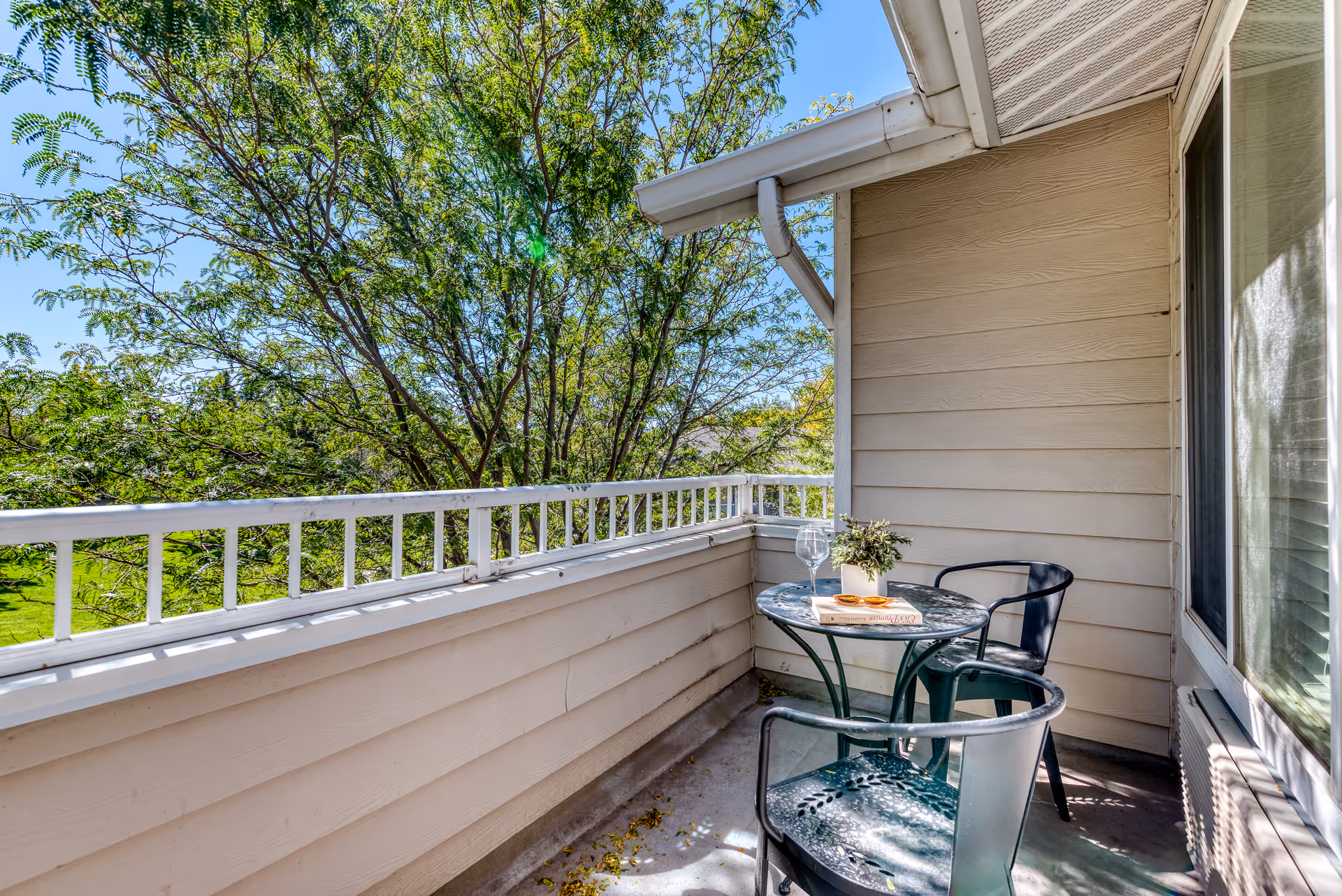 A small outdoor balcony area with a round glass table and two black metal chairs. On the table, there is a small potted plant, a book, and a wine glass. The balcony has beige siding and white railing, with green trees and blue sky visible in the background.