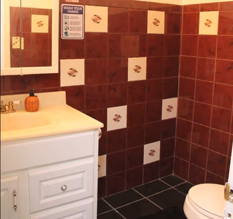 A bathroom with dark red and beige tiled walls featuring a geometric pattern. There is a white vanity with a sink and a small pumpkin decoration on the countertop. A mirror is mounted above the sink, and a sign reminding to wash hands is visible on the wall. A white toilet is partially visible on the right side.