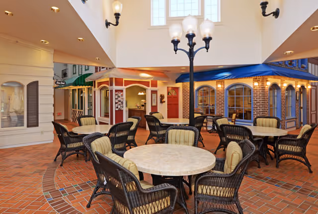 Indoor seating area with round tables and wicker chairs with cushions arranged on a brick floor. The space resembles a small indoor village with storefront-style facades, including a red door and windows with awnings. A tall black streetlamp with multiple lights stands in the center, and the ceiling has large windows allowing natural light.