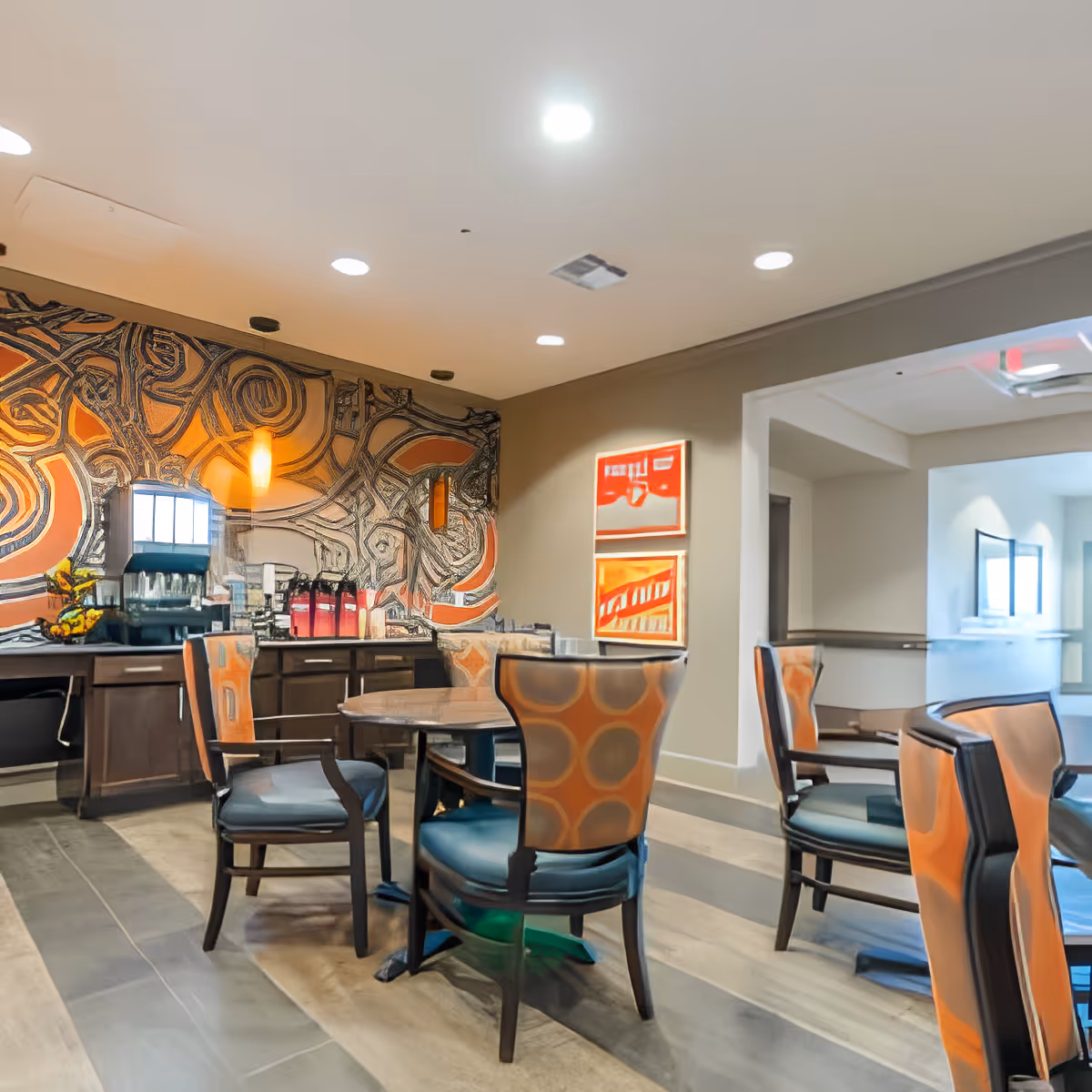 A dining area in a senior living facility with round tables and chairs featuring orange and blue upholstery. The back wall has an abstract mural with orange and black patterns. There is a counter with beverage dispensers and a coffee machine. Two framed abstract artworks hang on a beige wall near an open doorway leading to another room.