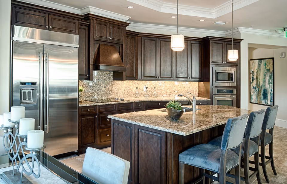 Spacious modern kitchen with a large granite island, dark wood cabinets, stainless steel refrigerator, and bar stools.