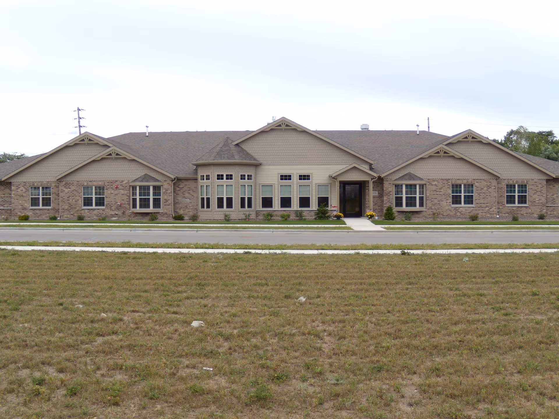 Front exterior view of a single-story senior living facility building with a combination of brick and beige siding, multiple windows, and a central entrance with a small covered porch. There is a grassy area and a sidewalk in front of the building.