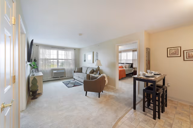 Bright living room with a sofa, armchair, wall-mounted TV, large window, and a small dining table with stools opening to a visible bedroom.