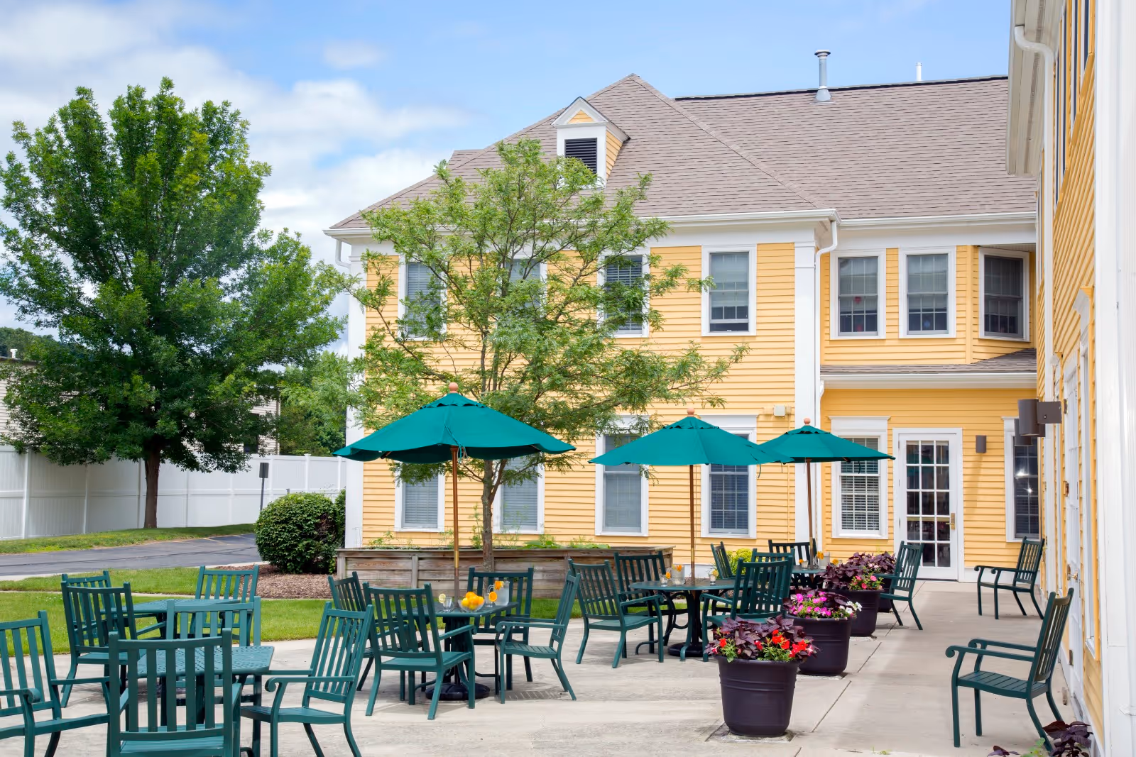 Outdoor patio area at a senior living facility with green tables and chairs, green umbrellas, potted flowers, and a yellow building with white trim in the background under a partly cloudy sky.
