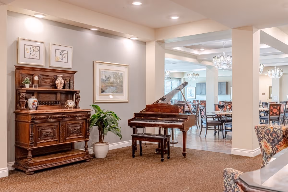 Interior view of a senior living facility featuring a wooden piano with a bench, an ornate wooden cabinet with decorative items, framed artwork on the walls, a potted plant, and a dining area with tables and chairs visible in the background under chandeliers.