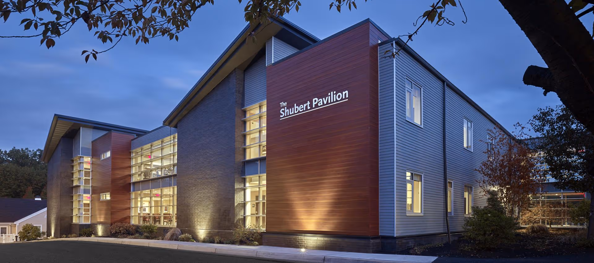 Exterior view of The Shubert Pavilion building at dusk, featuring modern architecture with large windows and illuminated walls, surrounded by trees and landscaping.