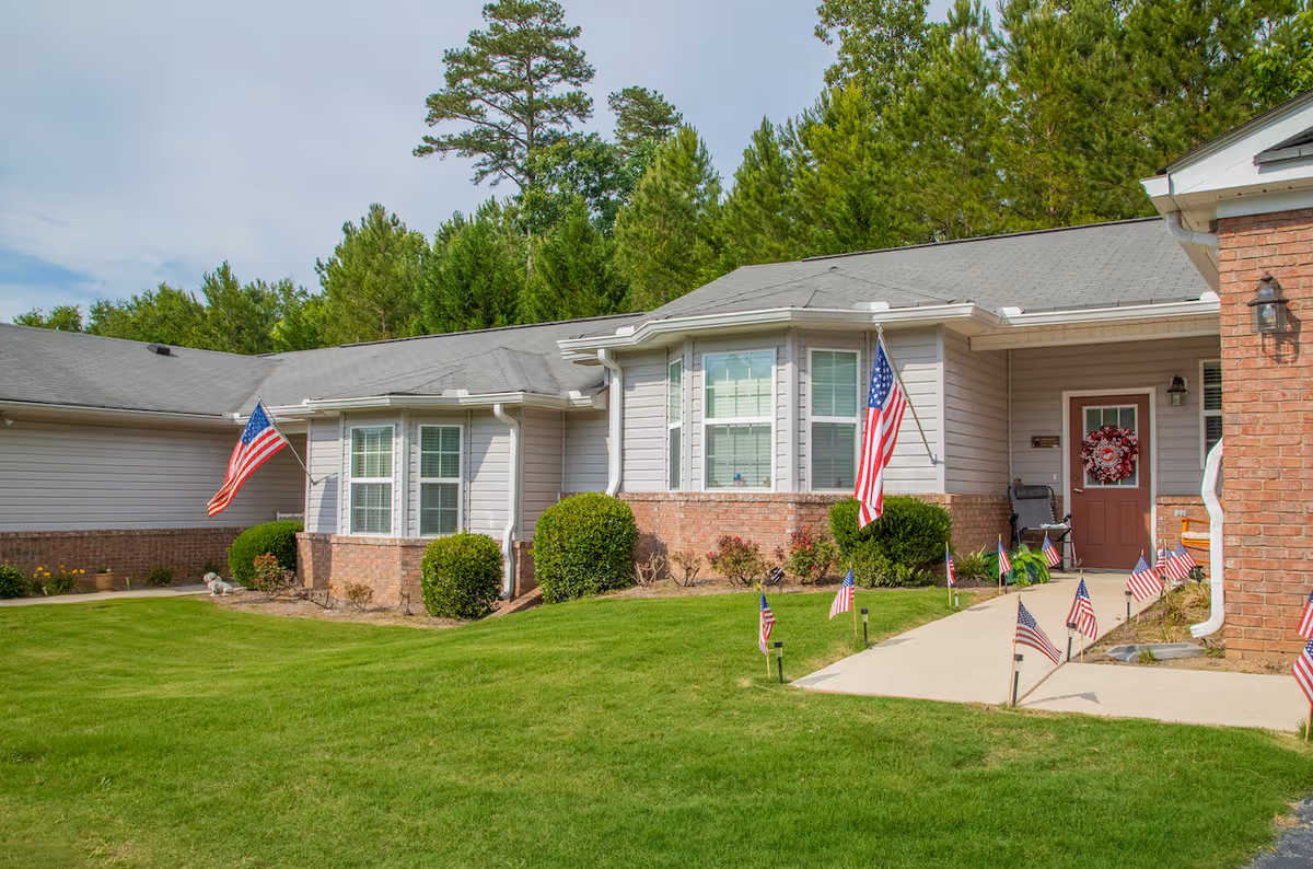 Exterior view of a single-story residential building with beige siding and brick accents, a gray roof, and multiple windows. The front entrance has a red door decorated with a wreath and a porch light. The walkway leading to the door is lined with small American flags, and two larger American flags are mounted on the building. The lawn is green and well-maintained, with bushes near the building and tall pine trees in the background under a partly cloudy sky.