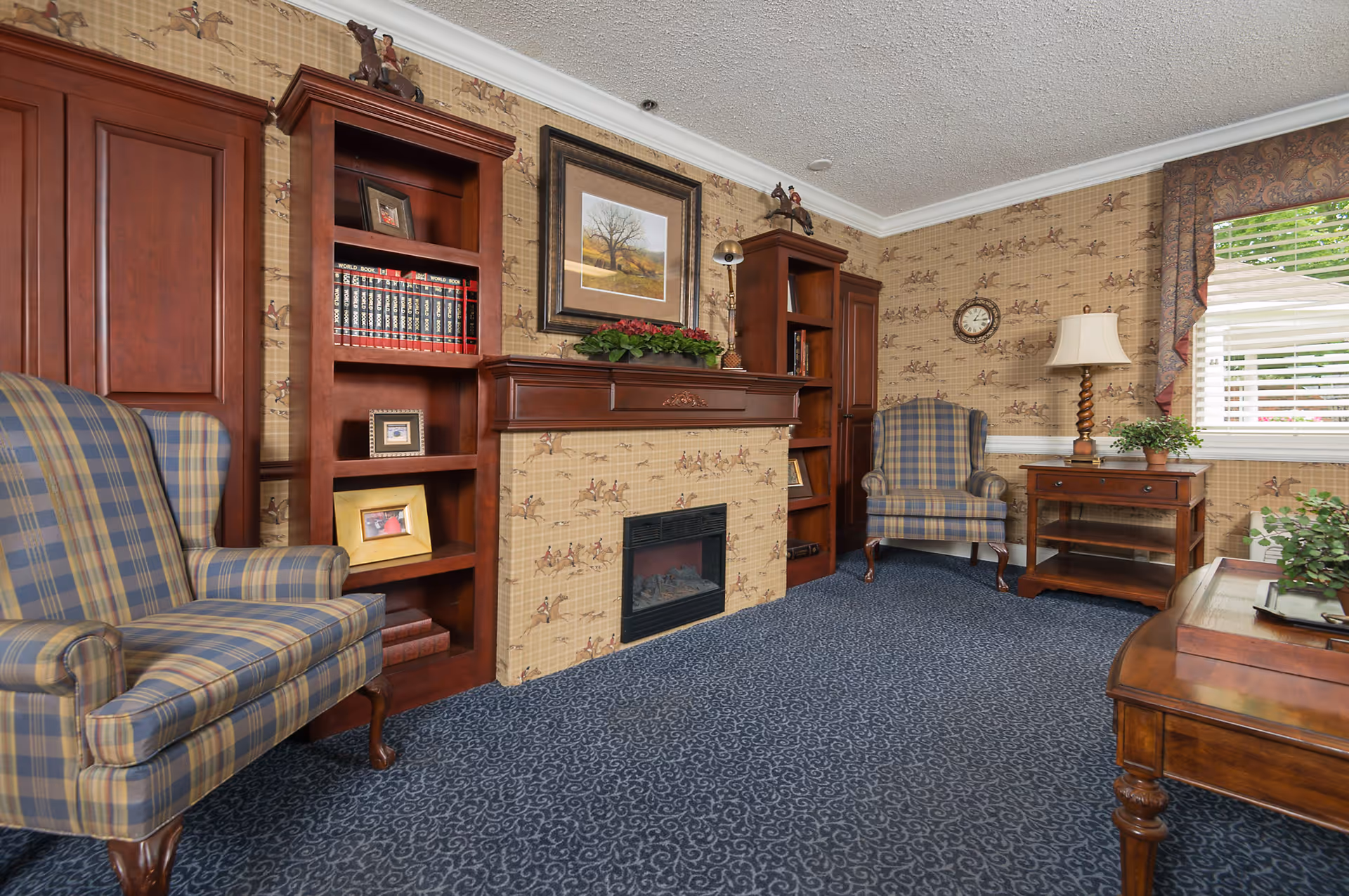 Well-appointed sitting room with two plaid wingback chairs, wooden bookshelves and a mantel fireplace.