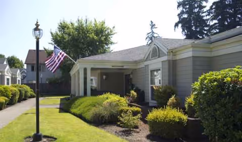 Single-story senior living facility exterior with lawn, shrubs, and an American flag.