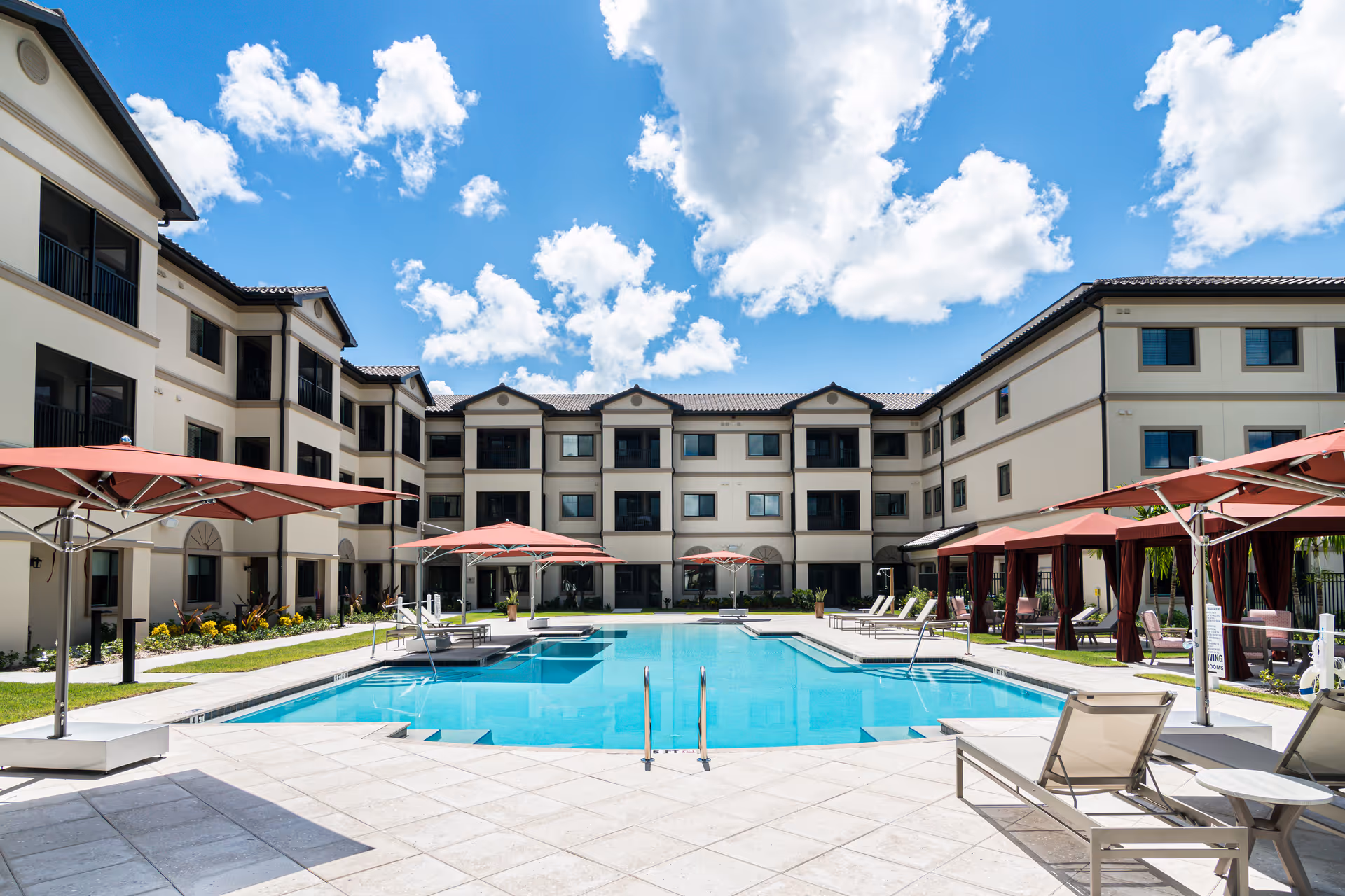 Outdoor swimming pool area at Discovery Village At Naples with lounge chairs, red umbrellas, and a three-story building surrounding the pool under a partly cloudy blue sky.
