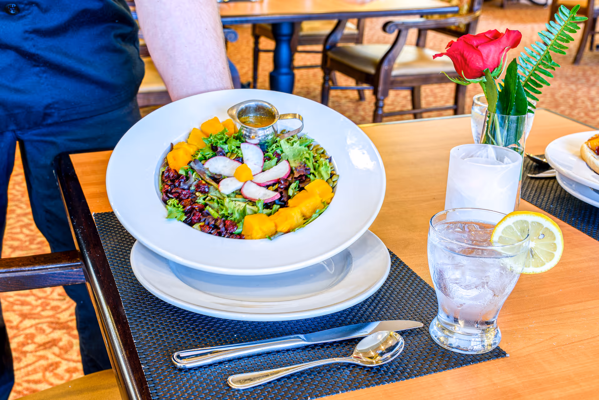 A fresh salad served on a white plate with mixed greens, sliced radishes, dried cranberries, and chunks of yellow fruit or vegetable, accompanied by a small metal container of dressing. The plate is placed on a black woven placemat on a wooden table with a glass of water garnished with a lemon slice, a red rose in a small vase, and silverware including a knife and spoon. A person wearing a dark blue shirt is partially visible holding the plate.