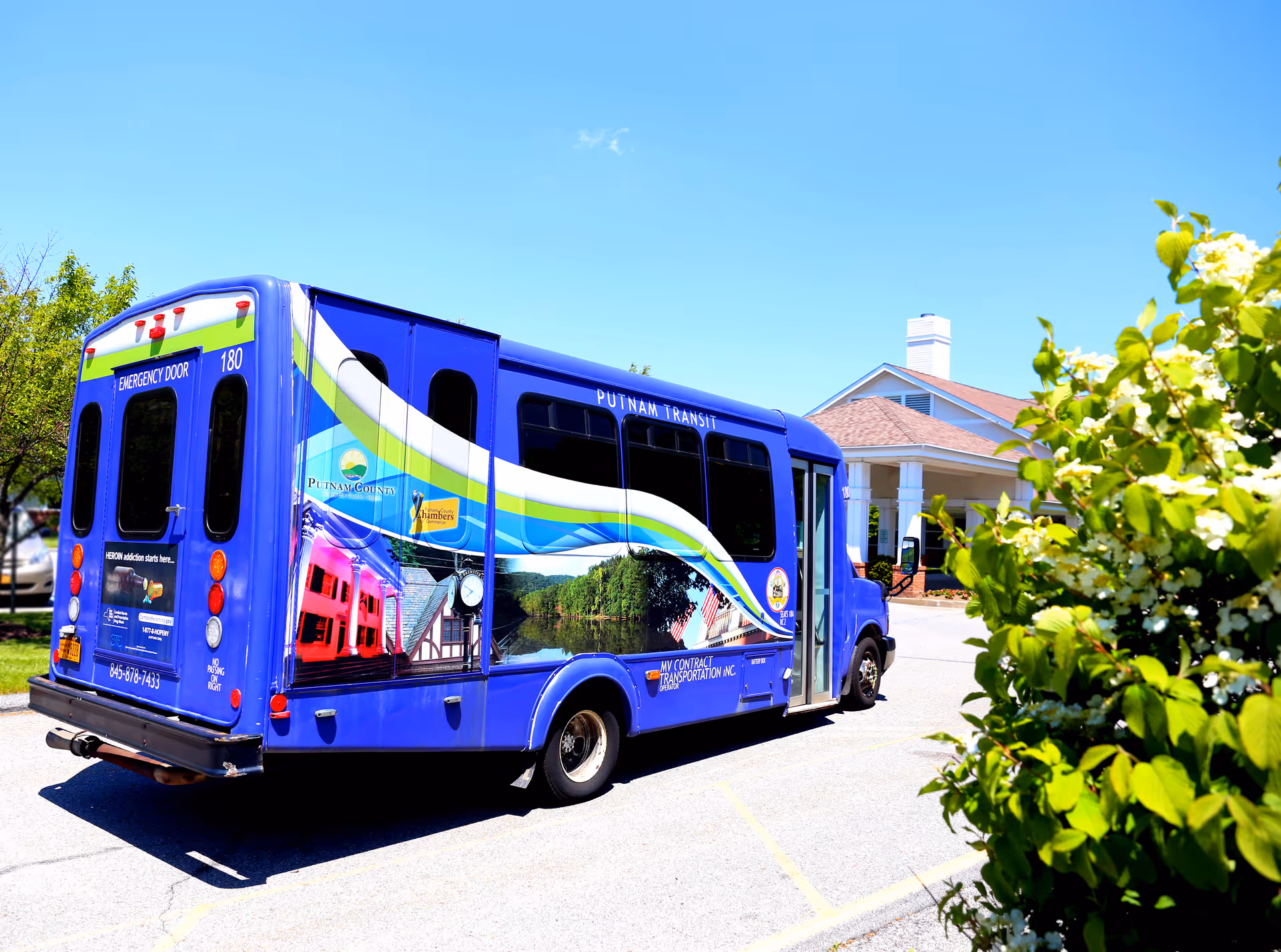 A blue Putnam Transit shuttle bus parked outside a building with a porch and chimney under a clear blue sky. Green bushes are visible in the foreground on the right side.