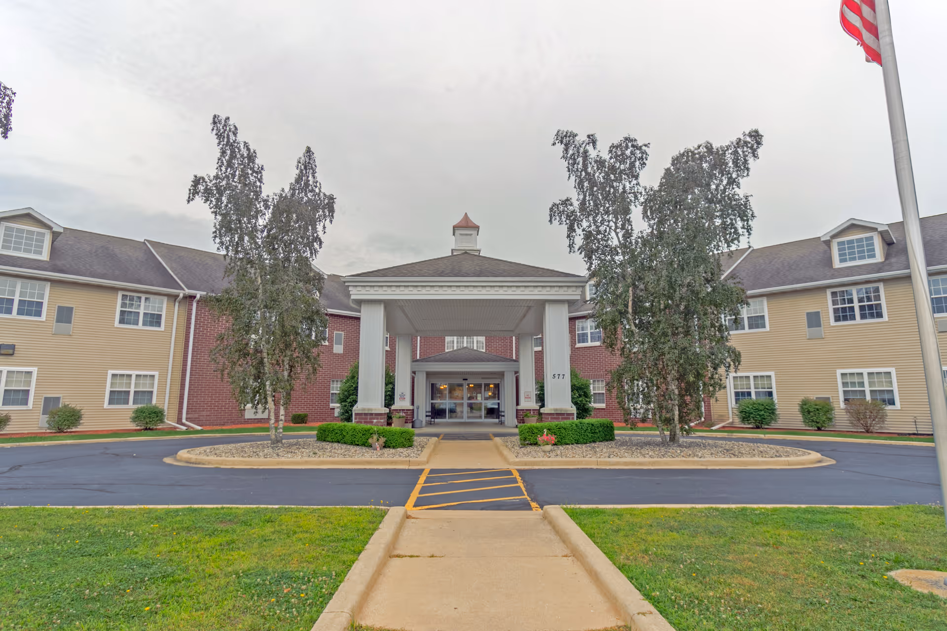 Front exterior view of Heritage Woods of Watseka building with a covered entrance supported by four white columns, two tall trees on either side, and an American flag on a flagpole to the right. The building has beige and red brick siding with multiple windows.