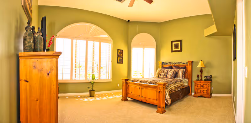 A spacious bedroom with green walls and beige carpet. The room features a wooden bed with patterned bedding, a matching wooden nightstand with a lamp, and a wooden dresser with decorative items on top. There are two large windows with white shutters allowing natural light to fill the room.