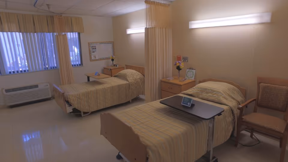 A nursing facility room with two single beds separated by a privacy curtain. Each bed has a beige quilted blanket and a small overbed table. There is a wooden nightstand with flowers and a small sign between the beds. A cushioned chair is placed next to one of the beds. The room has a window with vertical blinds and an air conditioning unit below it.