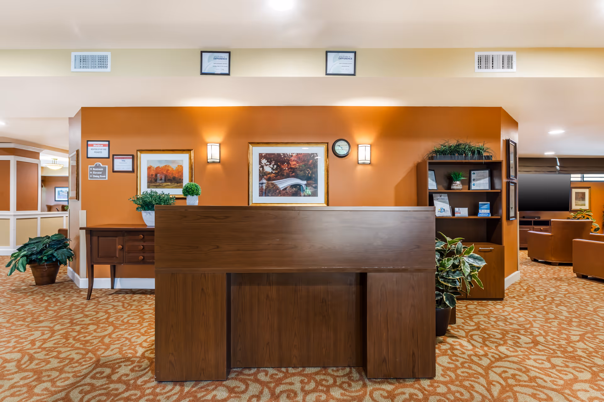 Reception desk area in a senior living facility with a wooden desk in the foreground, orange accent wall behind it featuring framed artwork, two wall sconces, a clock, and plants on either side. To the right, there is a bookshelf with informational pamphlets and more plants. The carpet has a patterned design in warm tones.