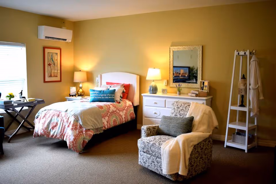 A cozy bedroom with a single bed covered in a patterned quilt and multiple pillows, including one with text. There is a bedside table with a lamp, a white dresser with a lamp and framed pictures, a decorative mirror on the wall, a patterned armchair with a throw blanket and pillow, a white shelving unit with a robe hanging, and a window letting in natural light.
