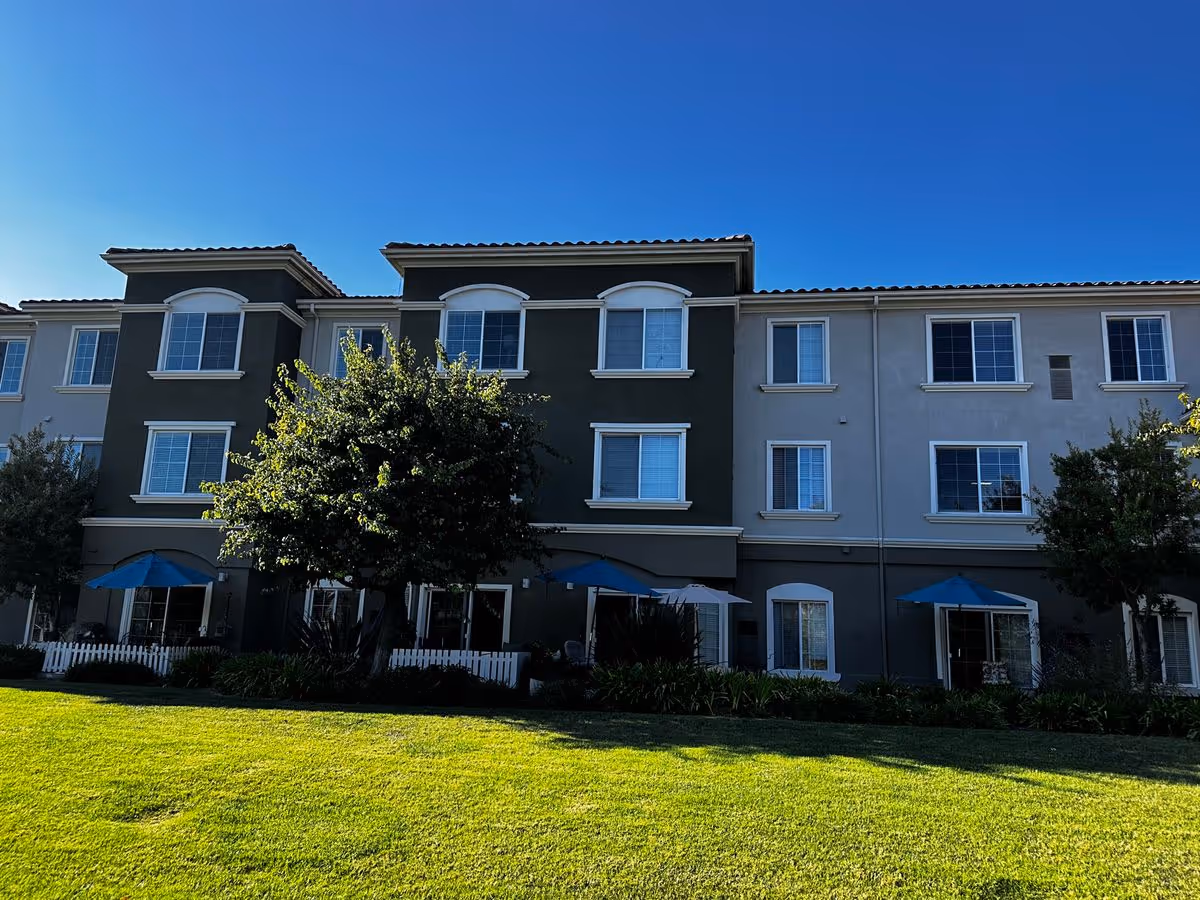 Front exterior of a three-story senior living building with a green lawn, trees, and blue patio umbrellas under a clear blue sky.