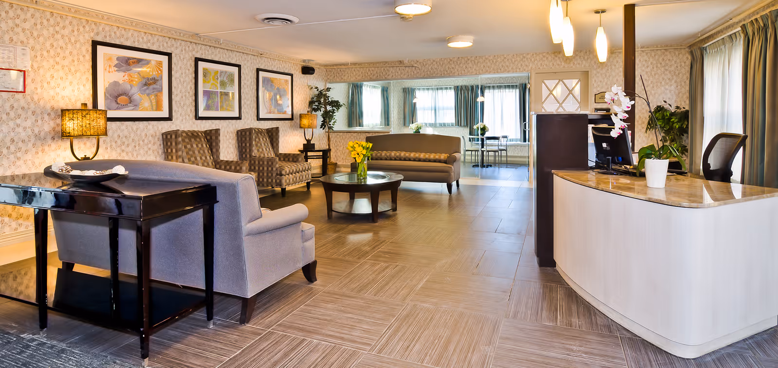 A spacious and well-lit senior living facility lobby with a curved reception desk on the right, a seating area with armchairs and a sofa, a round coffee table with a vase of yellow flowers, framed floral artwork on the wall, and large windows with curtains allowing natural light.
