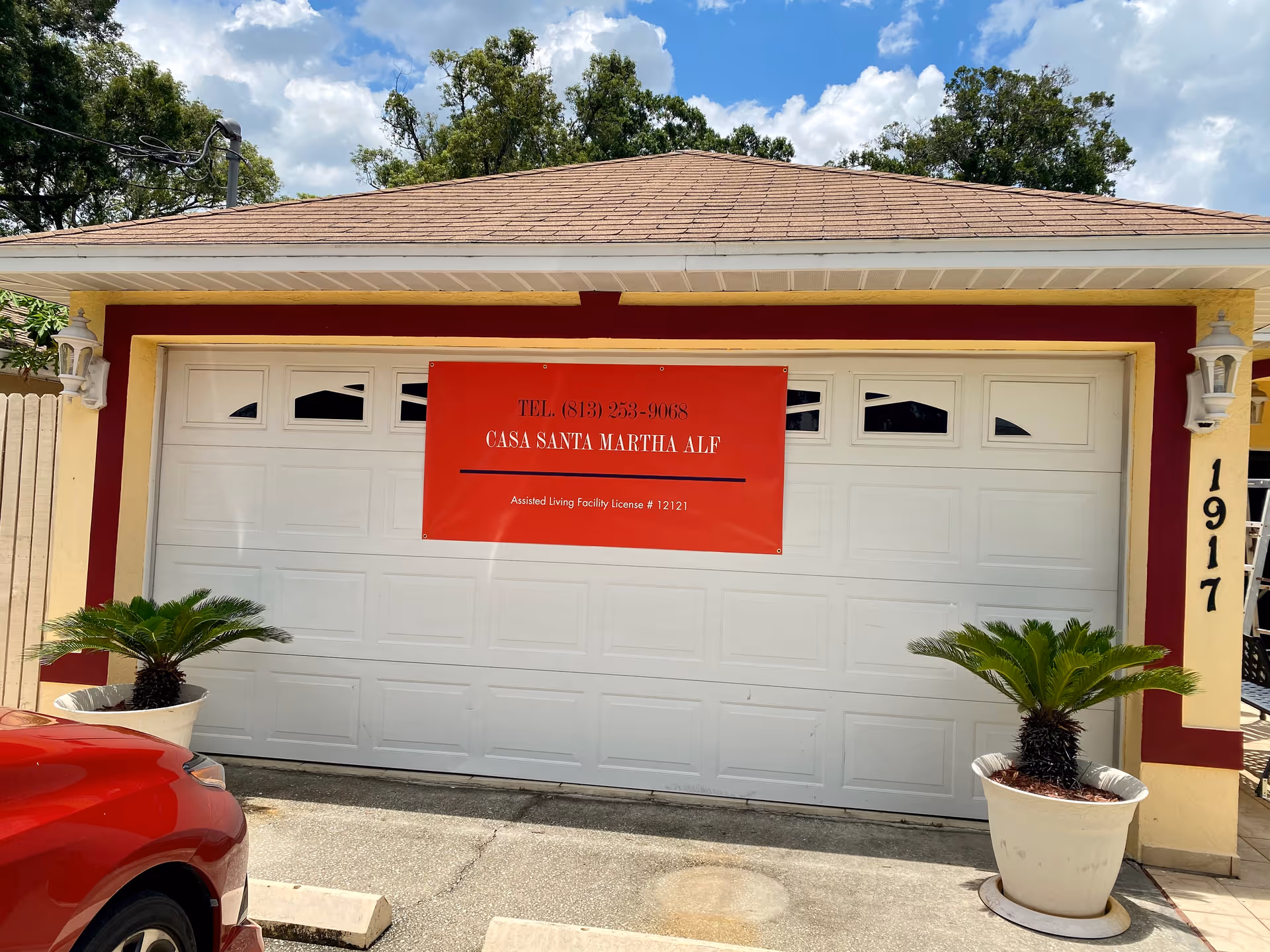 Front view of a garage door with a red banner displaying the text 'TEL. (813) 253-9068 CASA SANTA MARTHA ALF Assisted Living Facility License # 12121'. The garage is part of a building with beige and maroon trim, flanked by two potted plants. The building number 1917 is visible on the right side. A red car is partially visible in the foreground.