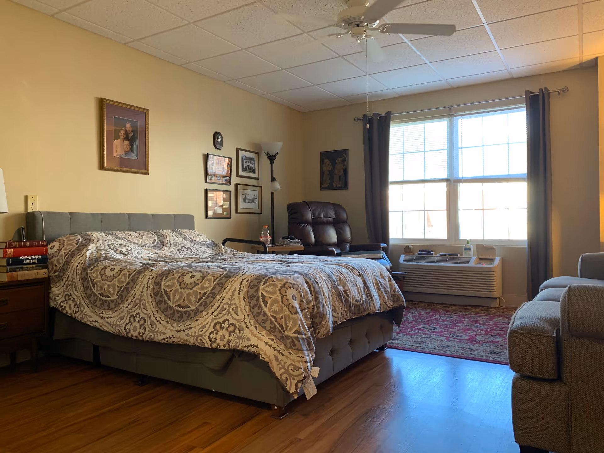 A cozy bedroom in an assisted living facility featuring a large bed with patterned bedding, a nightstand with books, a leather recliner chair, a floor lamp, framed pictures on the wall, a window with curtains, an air conditioning unit below the window, and a sofa partially visible on the right side.