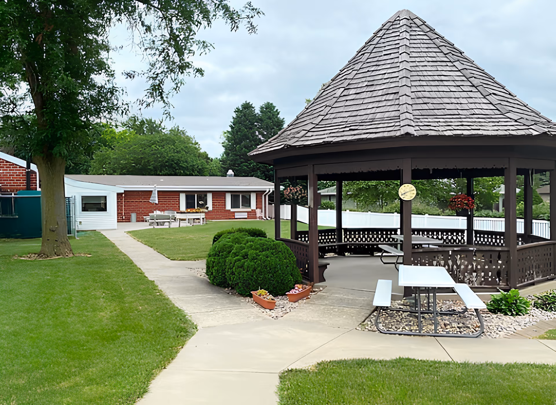 Outdoor area of Azria Health Rose Vista featuring a wooden gazebo with a shingled roof, a white picnic table with benches, green bushes, a tree, and a brick building in the background under a cloudy sky.