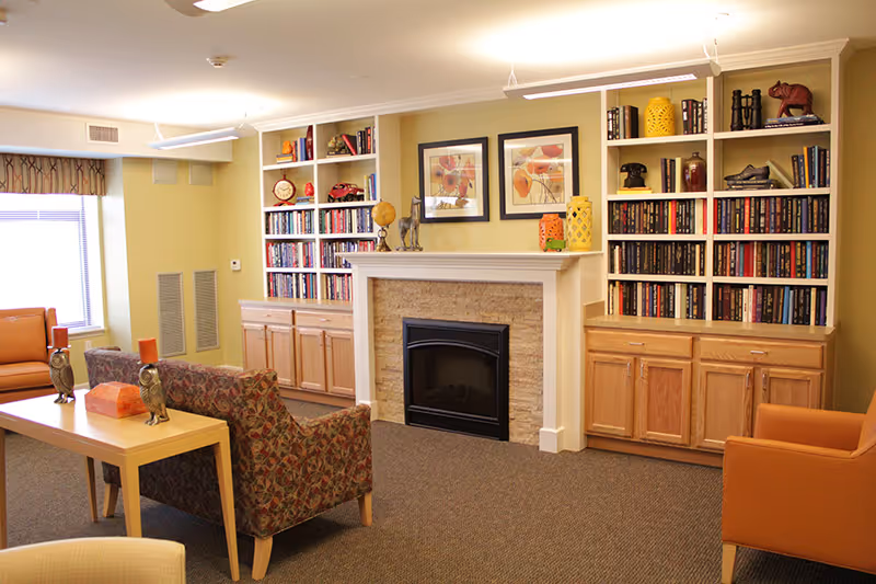 A cozy living room area in an assisted living facility featuring a fireplace with a stone surround and white mantel, flanked by built-in wooden bookshelves filled with books and decorative items. The room has patterned armchairs, an orange armchair, a wooden table with owl figurines and candles, and a window with blinds and a valance allowing natural light to enter.