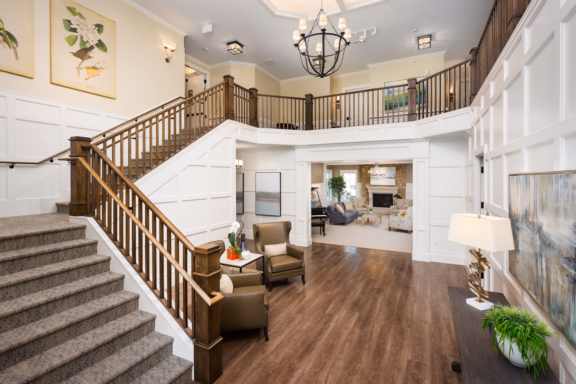 Spacious two-story senior living lobby with a central wooden staircase, seating area, chandelier, and a view into a fireplace lounge.