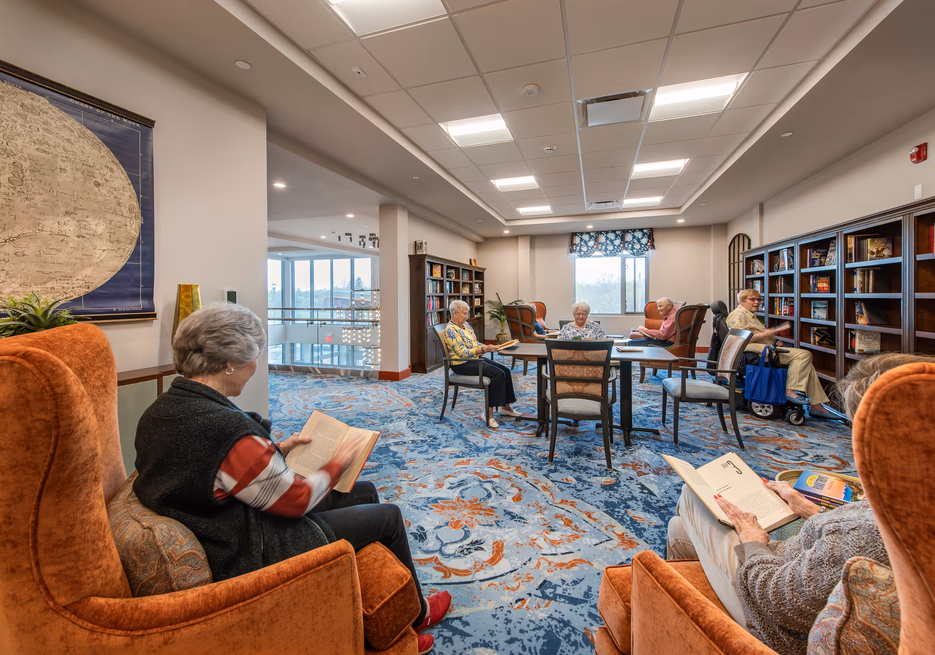 A cozy reading room in a senior living facility with several elderly people sitting in comfortable chairs and around a table, reading books. The room features a patterned blue and orange carpet, bookshelves filled with books, large windows letting in natural light, and warm orange armchairs.