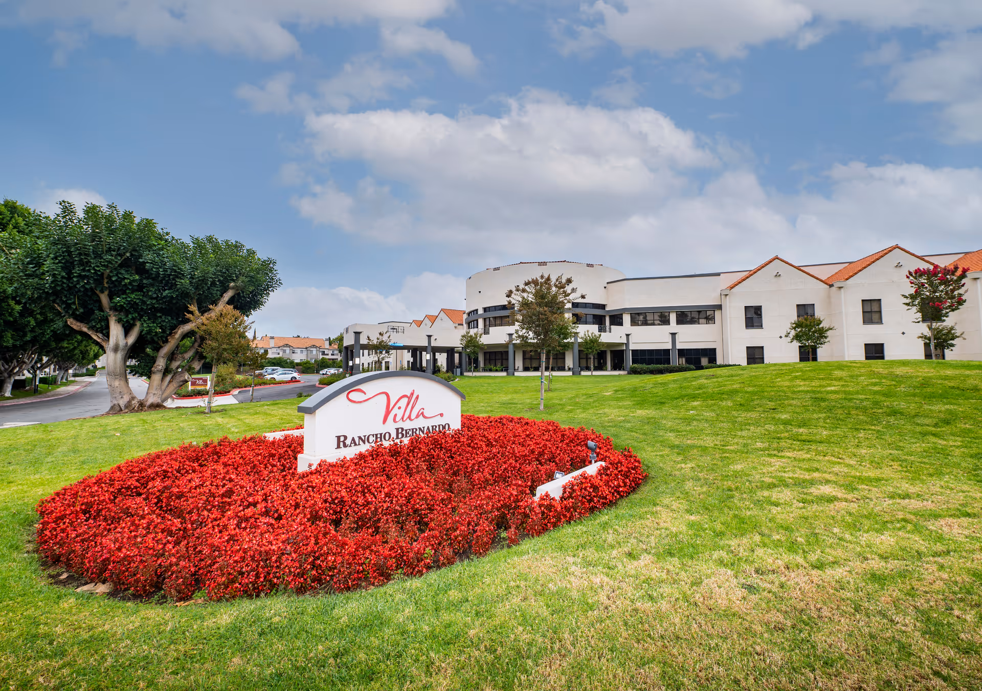 Exterior view of Villa Rancho Bernardo Care Center building with a manicured lawn and a flower bed with red flowers surrounding a sign displaying the facility's name. The building has white walls and a red-tiled roof under a partly cloudy sky.