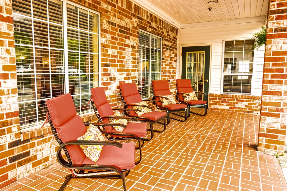 Covered outdoor patio area with five red cushioned chairs arranged in a row against a brick wall with large windows. Each chair has a decorative pillow. The floor is tiled with a brick pattern, and there is a green door with glass panes at the end of the patio.