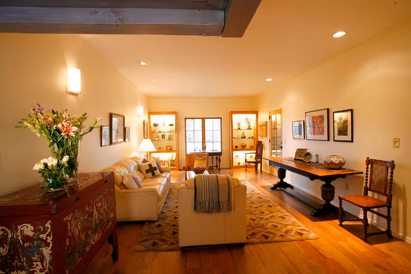 A warm living room with cream sofas, a patterned rug, wooden furniture, artwork on the walls and built-in illuminated shelves under a window.