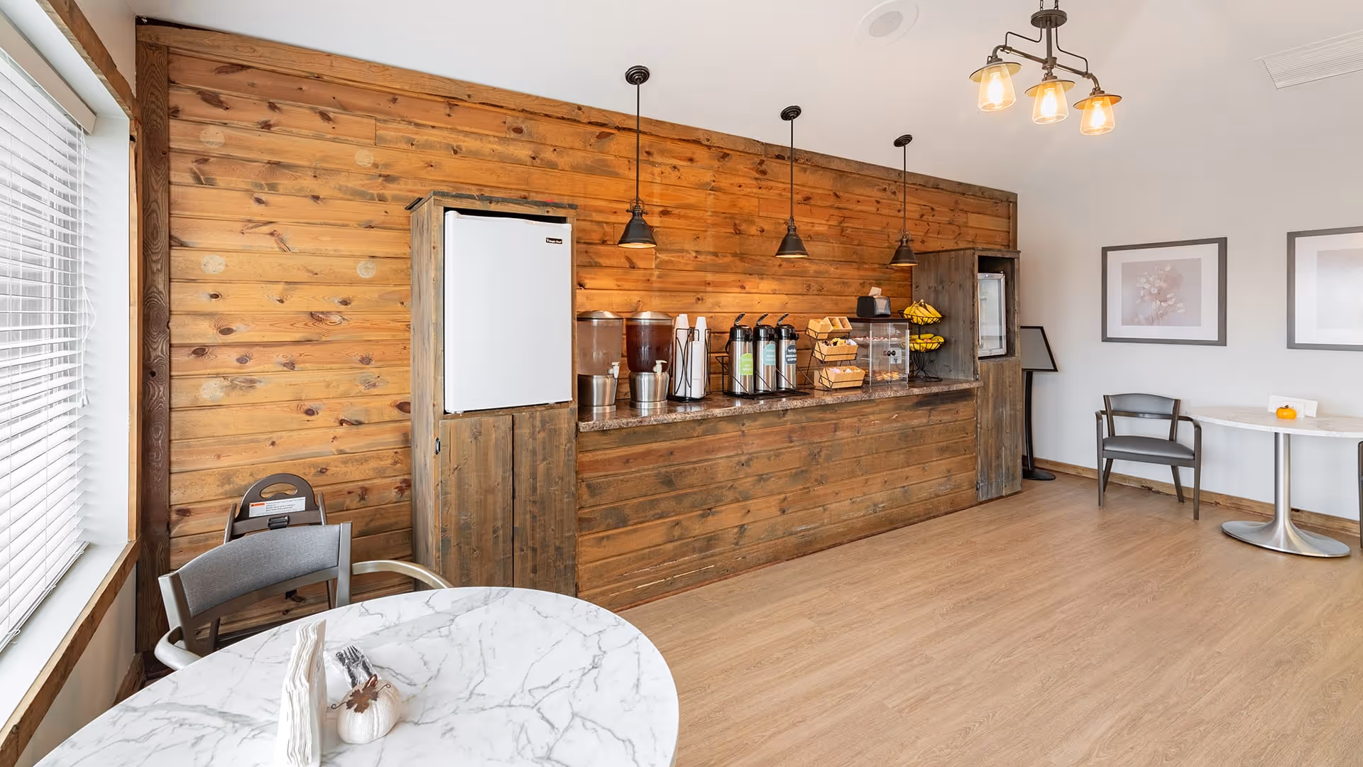 A cozy refreshment area with a wooden counter and wall, featuring a refrigerator, beverage dispensers, coffee urns, and baskets of snacks. There are two round marble tables with chairs near a window with blinds, and framed artwork on the white wall.