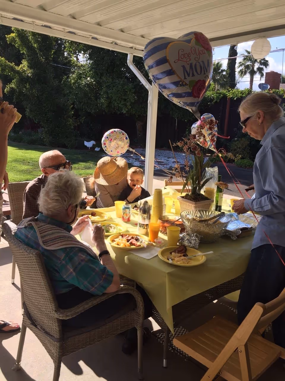A group of elderly people and a child gathered around a table outdoors under a covered patio. The table is covered with a yellow tablecloth and has plates of food, drinks, and a potted plant. There are balloons, including one heart-shaped balloon that says 'Love You Mom'. The setting appears to be a garden or backyard with green grass and trees in the background.